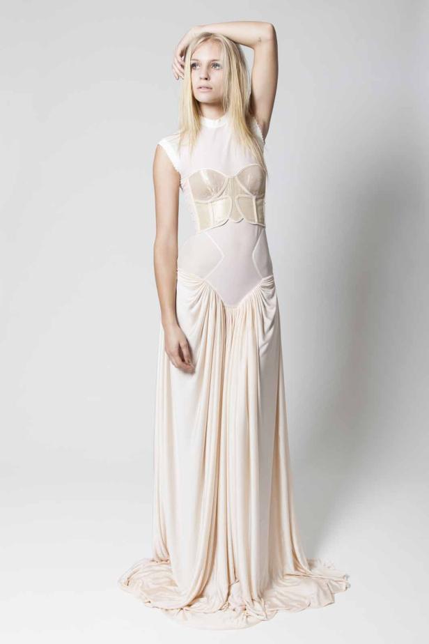 Eine blonde Frau in einem langen, hellen Kleid mit Korsett-Oberteil posiert vor einem weißen Hintergrund.