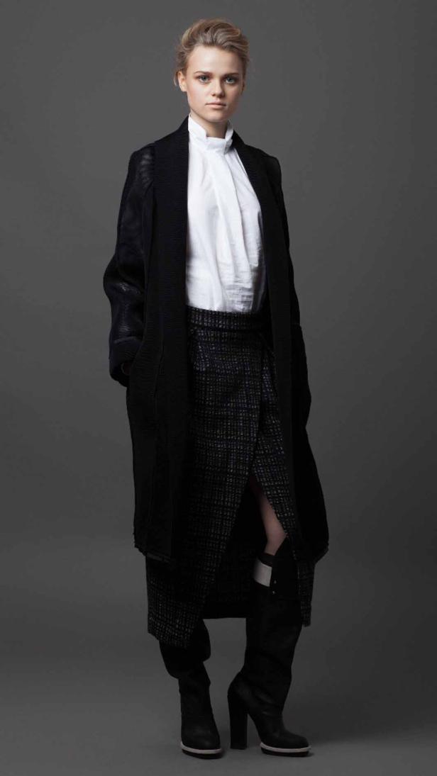 Eine Frau in einem langen, dunklen Mantel, einem weißen Hemd und einem gemusterten Rock mit Stiefeln.