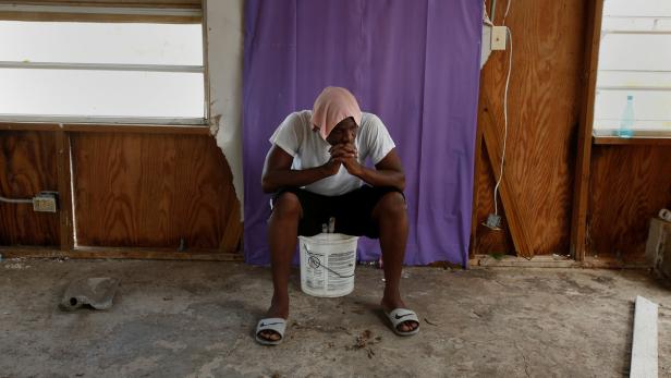 Ein Mann mit einem Handtuch auf dem Kopf sitzt nachdenklich auf einem Eimer in einem heruntergekommenen Raum.