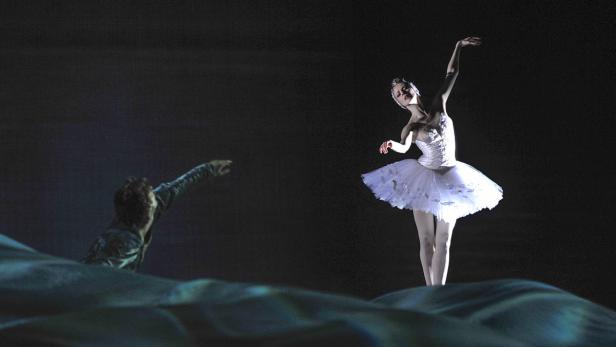 Eine Ballerina in einem weißen Tutu tanzt auf einer Bühne, während ein Mann ihr zusieht.