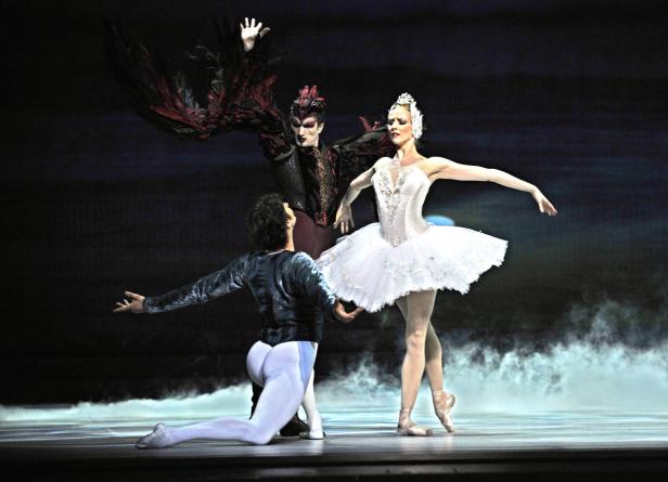 Eine Ballerina in Weiß tanzt mit zwei Tänzern auf einer Bühne.