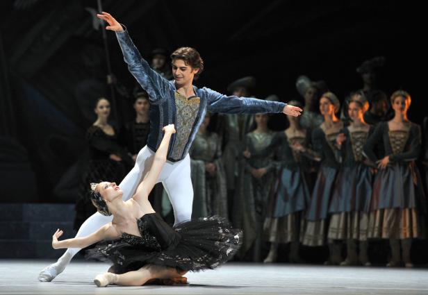 Eine Balletttänzerin in Schwarz wird von einem Tänzer in Blau gehalten, während andere Tänzer im Hintergrund stehen.
