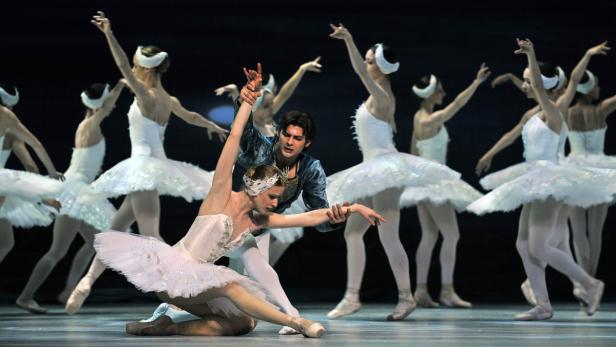 Eine Ballettaufführung mit einer Tänzerin im weißen Tutu, die von einem Tänzer gestützt wird, und weiteren Tänzerinnen im Hintergrund.