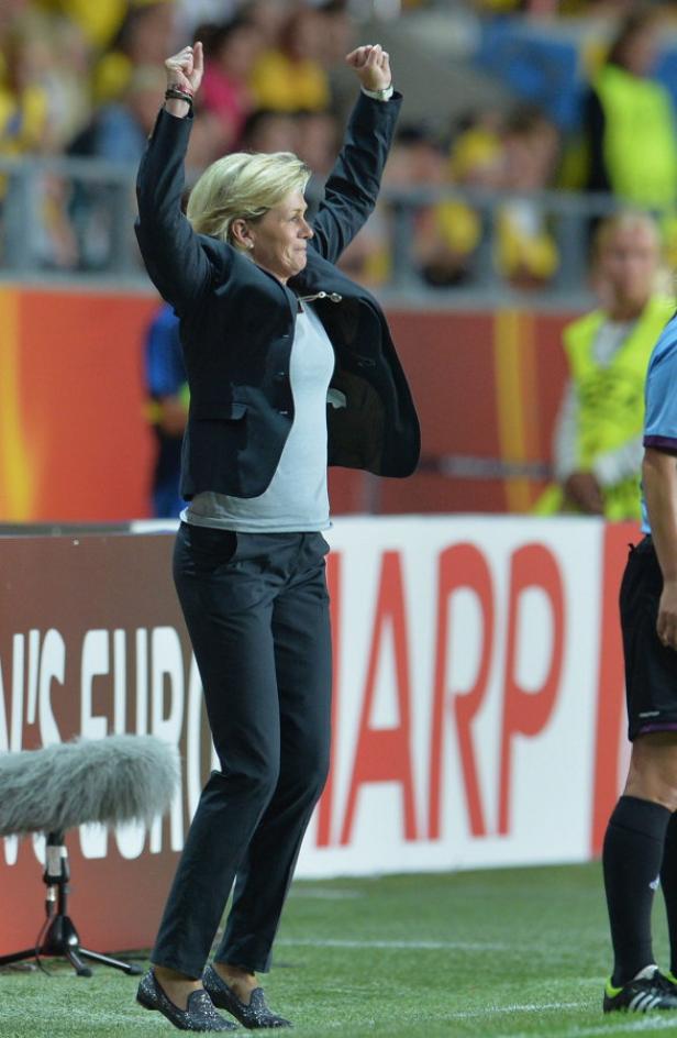 Eine Frau in einem Anzug jubelt mit erhobenen Armen auf einem Fußballfeld.