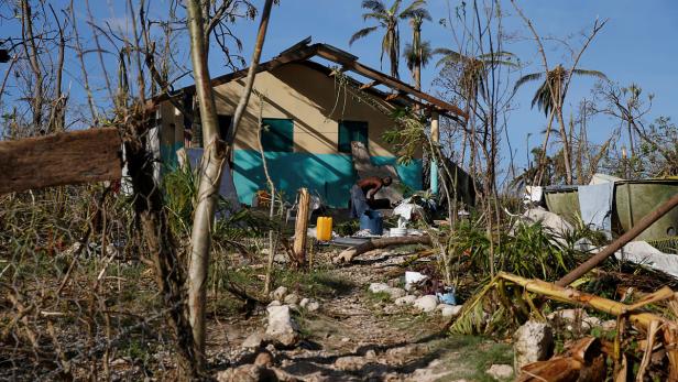 Ein Mann steht vor einem beschädigten Haus, umgeben von Trümmern und zerstörter Vegetation.