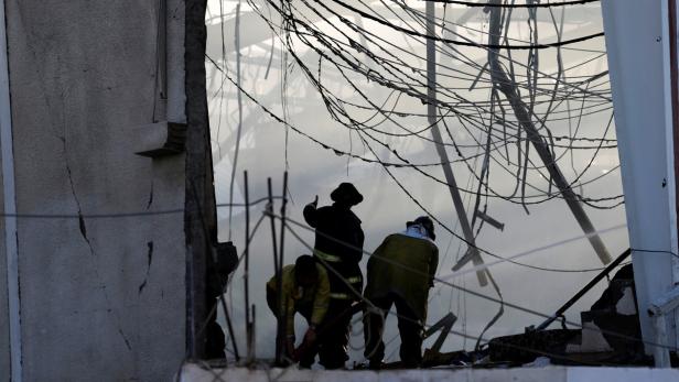 Drei Feuerwehrleute arbeiten im zerstörten Inneren eines Gebäudes, umgeben von Rauch und Trümmern.