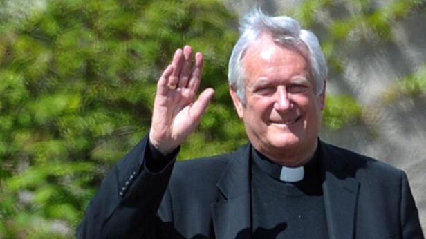 Ein Priester mit grauem Haar und schwarzem Talar winkt in die Kamera.