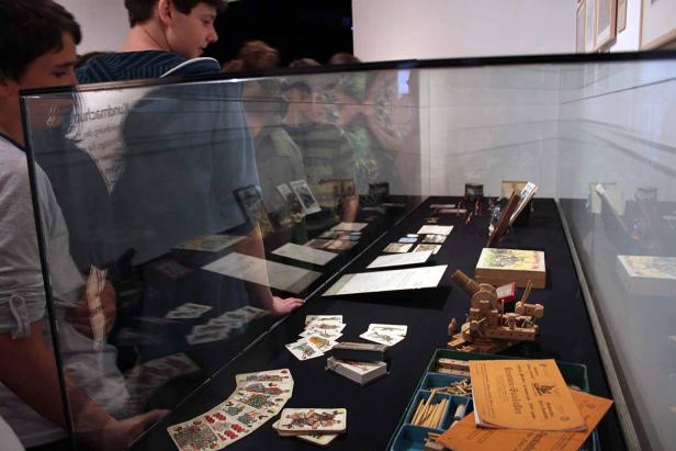 Besucher betrachten eine Ausstellung mit Spielkarten und einem Holzbaukasten.
