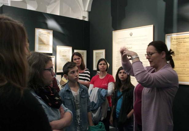 Eine Gruppe von Menschen hört in einem Museum einer Frau zu, die gestikuliert.