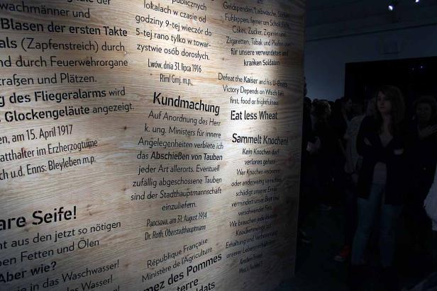 Eine Holzwand mit Texten in verschiedenen Sprachen, darunter Deutsch und Englisch, in einer Ausstellung.