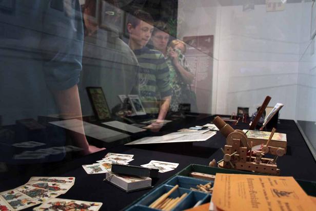 Besucher betrachten eine Ausstellung mit Spielkarten und einem Holzspielzeug-Kanonenmodell.