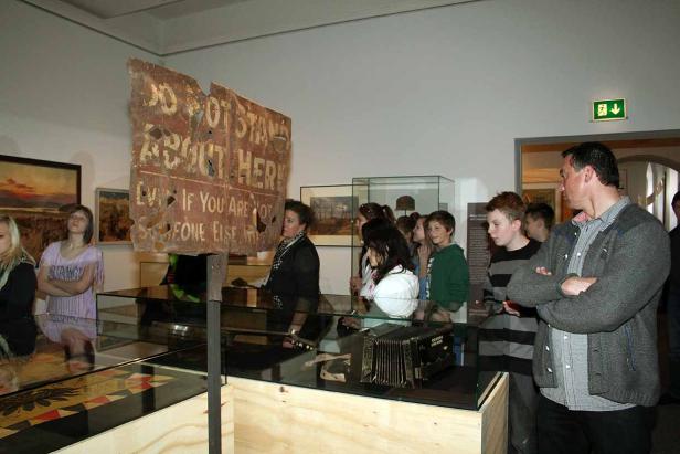 Eine Gruppe von Menschen betrachtet ein Schild mit der Aufschrift „Do not stand about here. Even if you are not alone elsewhere.“.
