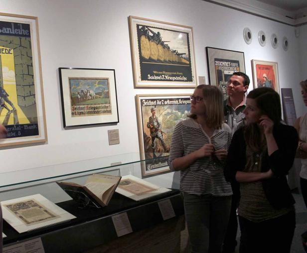Besucher betrachten eine Ausstellung mit historischen Kriegsanleihe-Plakaten.