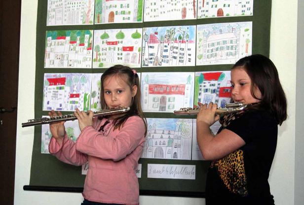 Zwei Mädchen spielen Flöte vor einer Wand mit Kinderzeichnungen von Häusern.