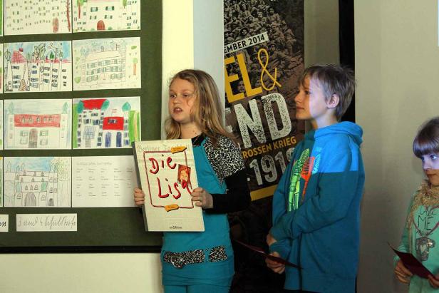 Ein Mädchen präsentiert das Buch „Die Lisa“ vor einer Wand mit Kinderzeichnungen.