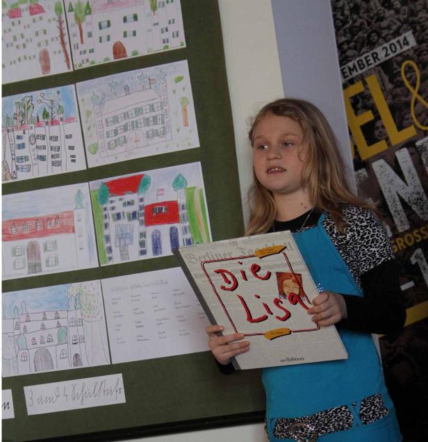 Ein Mädchen präsentiert das Buch „Die Lisa“ vor einer Wand mit Kinderzeichnungen.
