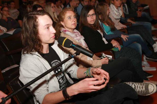 Ein junger Mann spricht bei einer Veranstaltung ins Mikrofon, umgeben von Zuhörern.