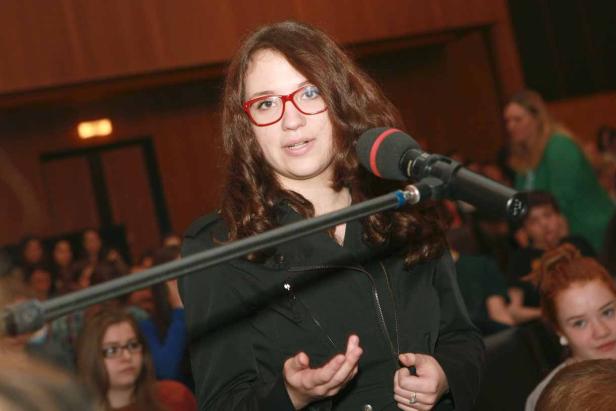 Eine junge Frau mit Brille spricht in ein Mikrofon vor Publikum.