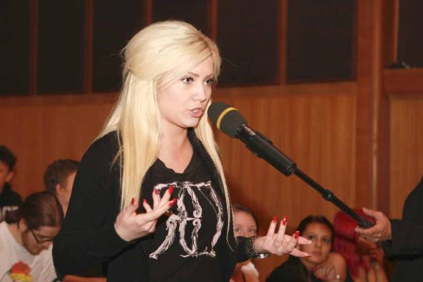 Eine blonde Frau spricht in ein Mikrofon vor einem Publikum.