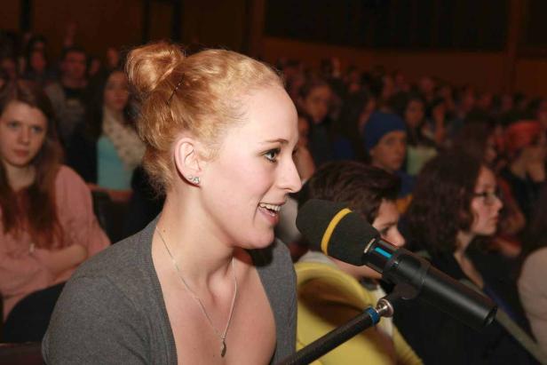 Eine junge Frau spricht vor einem Publikum in ein Mikrofon.