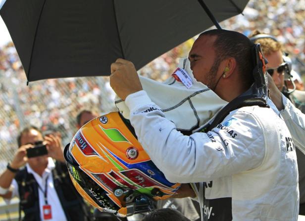 Lewis Hamilton wischt sich mit einem Tuch das Gesicht, während er einen Helm hält.