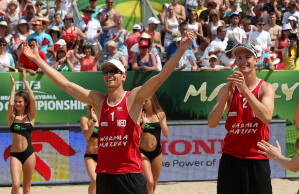 Zwei Beachvolleyballspieler jubeln am Strand, umgeben von Tänzern und Zuschauern.
