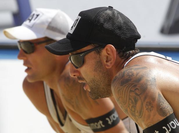 Zwei Beachvolleyballspieler mit Sonnenbrillen und Kappen erwarten den Aufschlag.