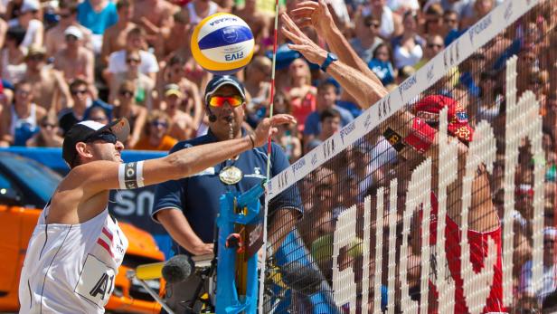 Ein Beachvolleyballspiel mit einem Schiedsrichter und zwei Spielern am Netz vor einer Zuschauermenge.