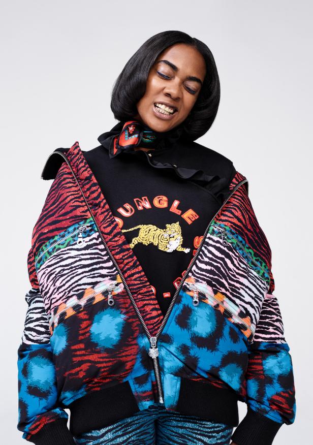 Eine Frau mit blauen Eyeliner trägt eine bunte Jacke mit Tiermustern über einem schwarzen Pullover mit dem Aufdruck „Jungle“.