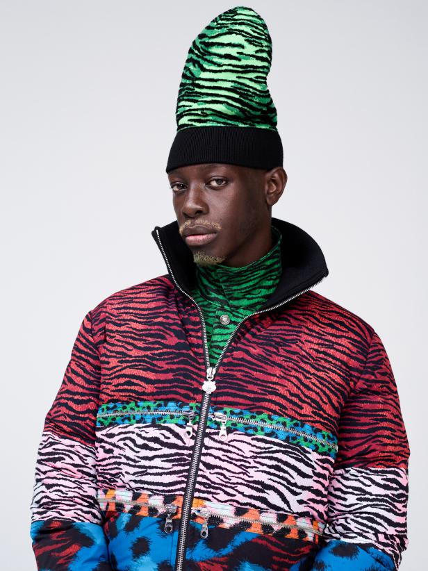 Ein Mann mit extravaganten Mustern auf Mütze, Rollkragenpullover und Jacke vor einem neutralen Hintergrund.