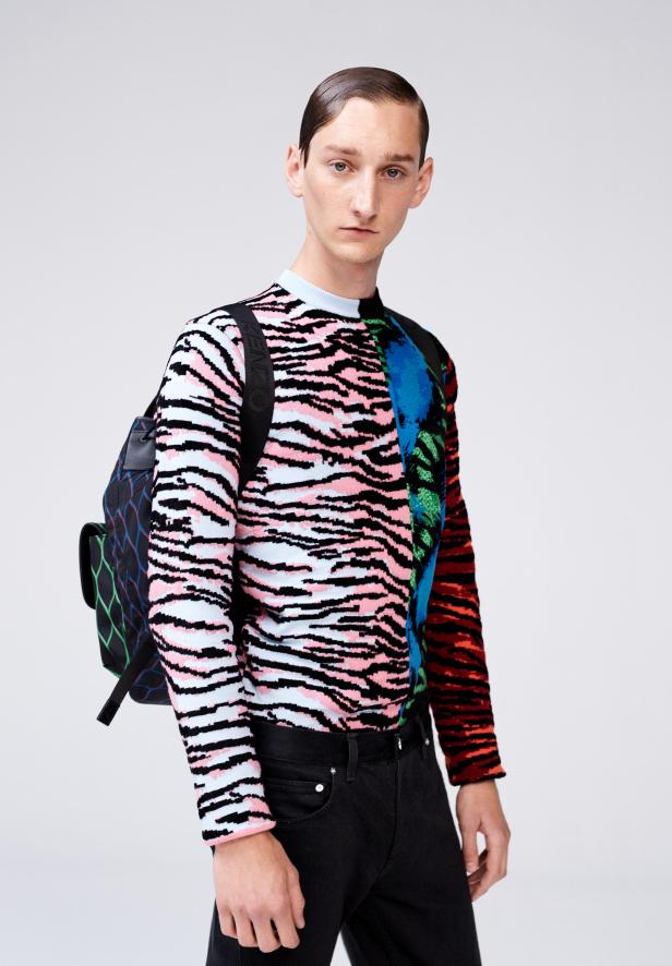 Ein Mann mit einem bunten Pullover und einem Rucksack vor einem neutralen Hintergrund.