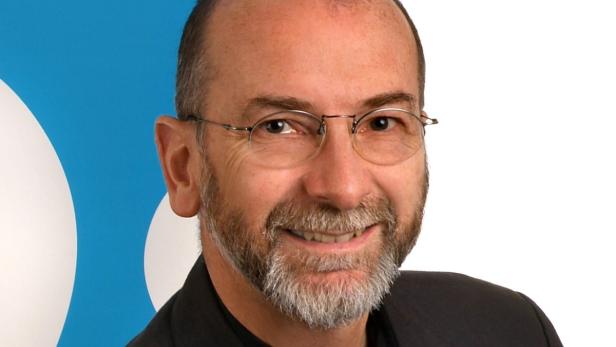 Porträt eines Mannes mit Brille und grauem Bart vor einem blauen Hintergrund.