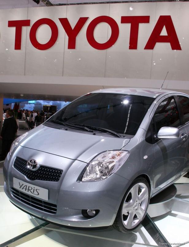 Ein silberner Toyota Yaris vor einem roten Toyota-Logo.