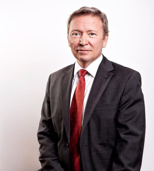 Ein Mann in einem dunklen Anzug und einer roten Krawatte blickt in die Kamera.