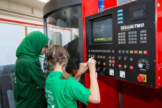 Ein Mädchen bedient eine CNC-Maschine, während eine Frau mit Kopftuch zusieht.