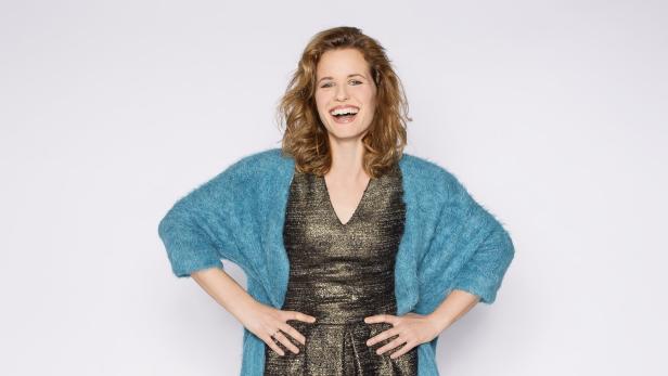 Eine lachende Frau mit einem türkisfarbenen Cardigan und einem goldenen Kleid posiert vor einem weißen Hintergrund.