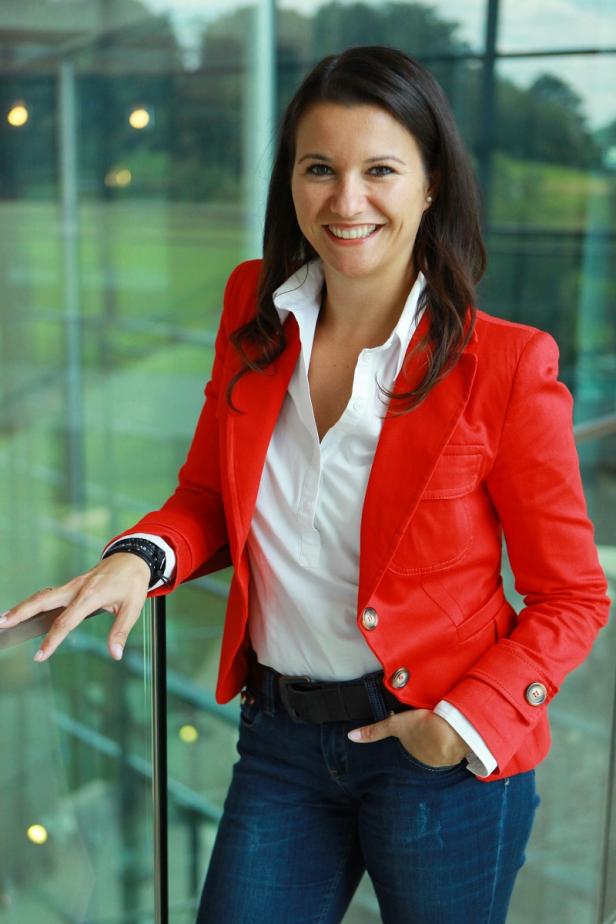 Eine Frau in rotem Blazer, weißer Bluse und Jeans lächelt in die Kamera.