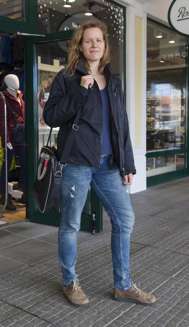 Eine Frau mit Jacke und Umhängetasche steht vor einem Geschäft.