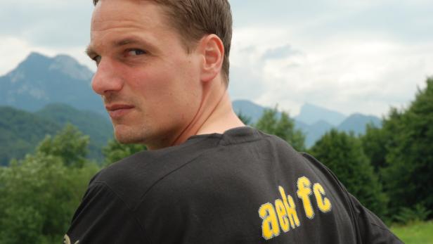 Ein Mann mit einem schwarzen T-Shirt und Bergen im Hintergrund blickt über seine Schulter.