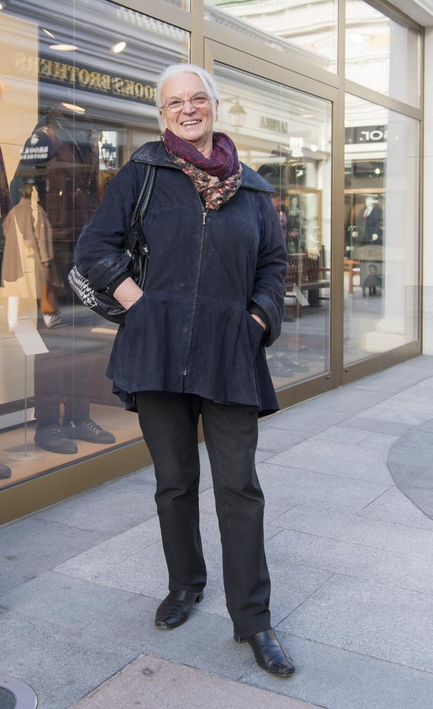 Eine ältere Frau mit Brille und Schal steht vor einem Schaufenster.