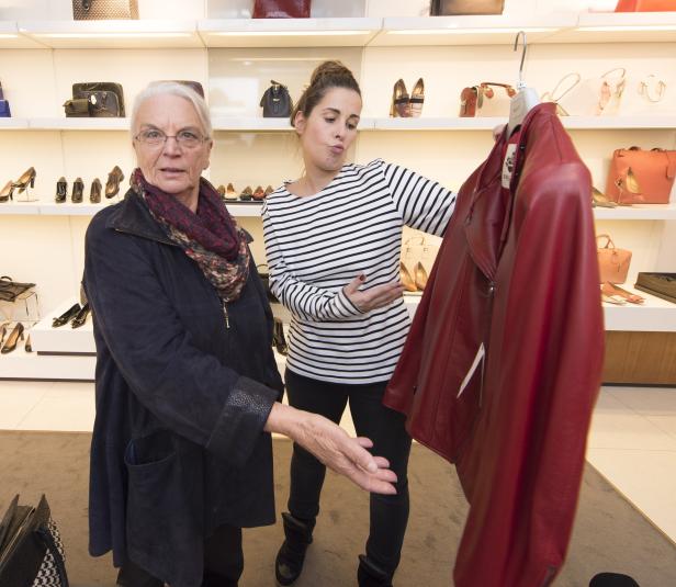 Eine ältere Frau betrachtet mit einer Verkäuferin eine rote Lederjacke in einem Schuhgeschäft.