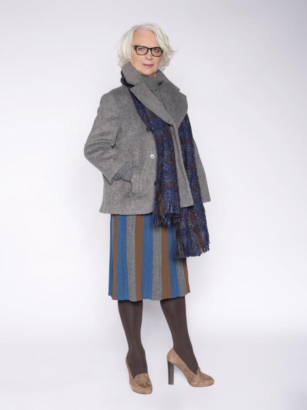 Eine ältere Frau mit Brille, grauem Mantel und gestreiftem Rock posiert vor einem weißen Hintergrund.