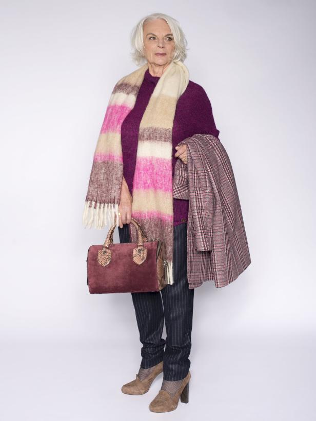 Eine ältere Frau mit grauem Haar, gekleidet in einen lila Pullover, Schal und Blazer, hält eine Handtasche.