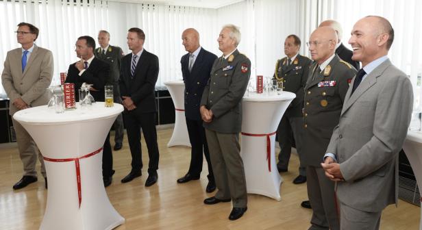 Eine Gruppe von Männern, einige in Uniform, steht in einem Raum mit Stehtischen.