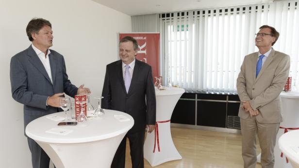 Drei Männer in Anzügen stehen in einem Raum mit Stehtischen und unterhalten sich.