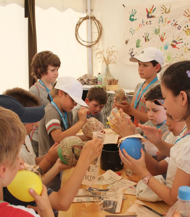 Eine Gruppe Kinder bastelt mit Pappmaché an einem Tisch, im Hintergrund ein Plakat mit der Aufschrift „Buntes Afrika“.