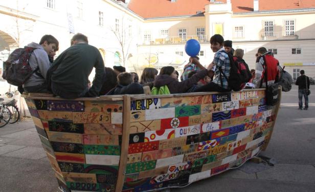 Eine Gruppe von Jugendlichen sitzt in einem bemalten Holzboot auf einem Platz.
