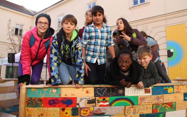 Eine Gruppe von Kindern steht hinter einer bemalten Holzwand mit dem Wort „Willkommen“.