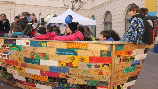 Kinder sitzen in einem bemalten Holzboot mit Aufschriften wie „Refugees Welcome“.
