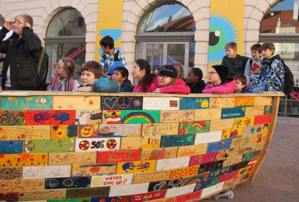 Eine Gruppe von Kindern steht in einem bemalten Holzboot mit Aufschriften wie „Refugees“ und „Zukunft“.